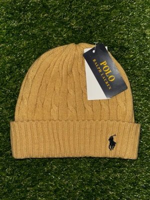 Ralph Lauren kabelstickad mössa - Snygg beige kabelstickad mössa från Polo Ralph Lauren med broderad logga i mörkblå framtill. Mjuk och varm, perfekt för kyliga dagar. Stilren design som passar till det mesta.