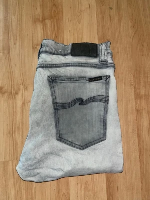 Nudie Jeans - Säljer ett par ljusgrå skinny jeans från Nudie Jeans med svarta kontrastsömmar. Midja 39cm, längd 109cm. OBS jeansen har 2 defekter: tyget är lite förtunnat samt slitna längst ned vid benöppningen.  Ifall det inte finns någon bild i annonsen där jag bär plaggen jag säljer så är det för att jag ej kan ha storleken heller passformen, så kolla måtten för storlek och googla på modellnamnet för passform 😉 s6,1