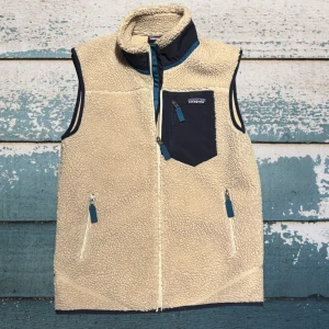 Beige fleeceväst från Patagonia - Mysig beige fleeceväst från Patagonia med marinblå detaljer. Västen har hög krage, dragkedja framtill, två sidofickor och en bröstficka med dragkedja. Perfekt lager på lager och riktigt skön att bära under kyliga dagar.