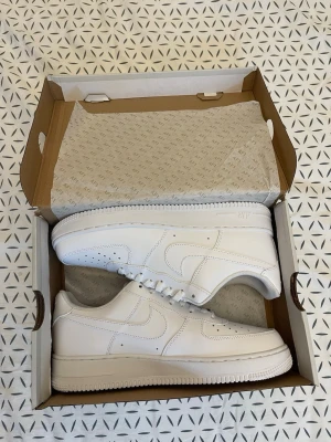 Nike Air Force 1 vita sneakers - Klassiska helvita Nike Air Force 1 sneakers i lågt utförande. Skorna har en stilren design med perforerade detaljer på tån, vit sula och ikoniska swoosh-loggan på sidan. Tillverkade i slätt läder med vita skosnören och diskret Air-märkning på sulan.