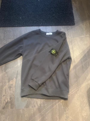 Stone Island tröja  - Säljer en stilren mörkgrå sweatshirt från Stone Island med den klassiska logotypen på ärmen. Tröjan har rund halsringning, ribbade muddar och är gjord i mjukt bomullsmaterial. Perfekt för dig som gillar streetwear och vill ha något bekvämt och snyggt.