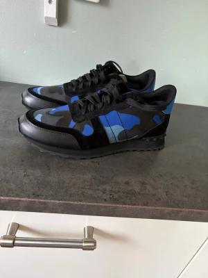 Valentino rockerrunners blå/svarta camo sneakers - Snygga sneakers från Valentino med blått och svart camo-mönster. Skorna har svarta detaljer i mocka och läder, samt svarta skosnören. Märkeslogga på plösen och en klassisk låg siluett. Perfekta för dig som vill sticka ut med stil. Dessa skor är sprillans nya från Valentino däremot för små vilket är orsaken till försäljningen.