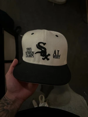 Vit och svart Chicago White Sox keps - Snygg vit och svart keps från New Era med Chicago White Sox-logga framtill. Broderad text '1993 Division Champs' och 'AL West' på framsidan samt MLB-logga bak. Klassisk flat peak och strukturerad passform, perfekt för dig som gillar sportig stil. Aldrig använd