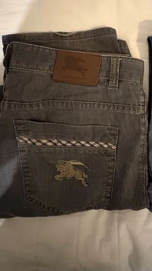 Gråa Burberry jeans med brodyr - Snygga gråa jeans från Burberry med klassisk broderad logga på bakfickan och rutigt band ovanför fickan. Jeansen har raka ben och detaljer i brunt läder vid linningen. Perfekta för dig som gillar exklusiva och stilrena plagg. Storlek 34 W 32 L kom med frågor och bud allt är intressant!