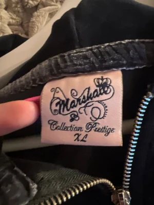 Svart velourhoodie Marshall XL - Snygg svart hoodie i mjuk velour från Marshall Collection Prestige. Dragkedja framtill, huva med snören och stor broderad logga i rött och vitt på bröstet. Den är i XL men mer som en S den är helt oanvänd. 