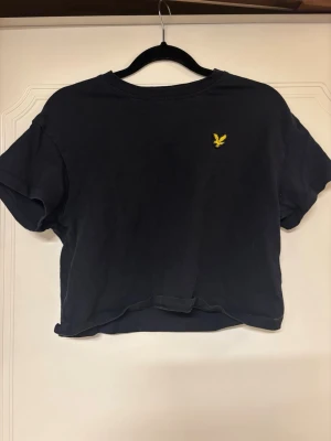 Svart croppad topp från Lyle & Scott - Svart croppad topp från Lyle & Scott med korta ärmar och gul broderad logga på bröstet. Perfekt till jeans eller kjol för en chill och trendig look.