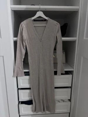 Beige ribbad långklänning med knappar - Långklänning  i beige ribbat material. Klänningen har v-ringning, långa ärmar och knappar hela vägen framtill. Tyvärr har knappen längst ner försvunnit men inget som märks tydligt 