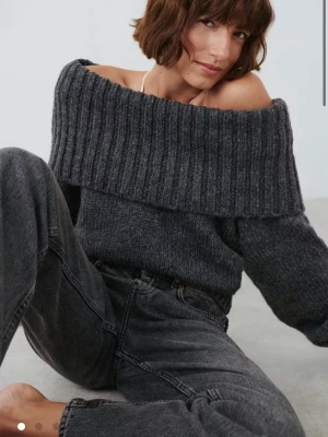 Mörkgrå off shoulder stickad tröja - Mysig mörkgrå stickad tröja med bred ribbad off shoulder-krage och långa ärmar. Tröjan har en avslappnad passform och är tillverkad i ett mjukt, grovstickat material som passar perfekt för kyliga dagar.