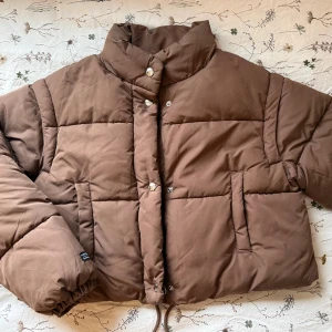 Brun pufferjacka - Säljer en brun pufferjacka med krage och dragkedja samt tryckknappar framtill. Jackan har två sidofickor och en boxig, croppad passform. Ena innerfickan har sprätts upp men annars som helt ny! (Trots att ena innerfickan är trasig så är det inget som märks, inget kommer trilla ut)🤎