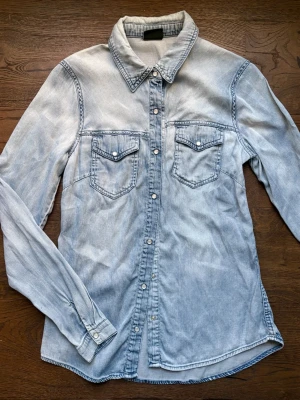 Ljusblå jeansskjorta från VILA - Ljusblå jeansskjorta från VILA med långa ärmar och tryckknappar framtill. 