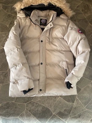 Ljusbeige dunjacka från Canada Goose - Snygg ljusbeige dunjacka från Canada Goose med klassisk patch på ärmen och stor huva med fuskpäls. Jackan har dragkedja och knappar framtill samt ribbade muddar vid ärmsluten. Perfekt för kalla vinterdagar.