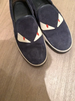 Fendi slip-on sneakers med ögon - Mörkblå slip-on sneakers från Fendi i mocka med ikoniska monsterögon i vitt och rött framtill. Skorna har vita sulor och coola metallspikar bak på hälen. Perfekt för dig som vill sticka ut med en unik och edgy stil.