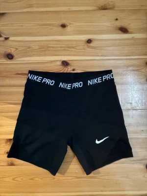 Svarta Nike Pro träningstights shorts - Säljer ett par svarta Nike Pro shorts med tight passform och elastisk midja där det står 'NIKE PRO' runtom. Perfekta för träning eller sport. Materialet är stretchigt och andas, med en liten vit Nike-logga på benet.