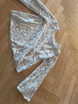 Vit spetsblus med v-ringning - Säljer en vit blus i spets med stora blommönster och v-ringning. Toppen har långa ärmar och dekorativa band i ryggen. Perfekt för dig som gillar romantisk och trendig stil.