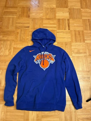 Blå New York Knicks hoodie från Nike - Snygg blå hoodie från Nike med New York Knicks-logga framtill. Klassisk huva med dragsko och stor magficka. Perfekt för basketfans som vill sticka ut med en sportig och bekväm stil. Materialet är mjukt och skönt, och trycket är i orange, vitt och grått.