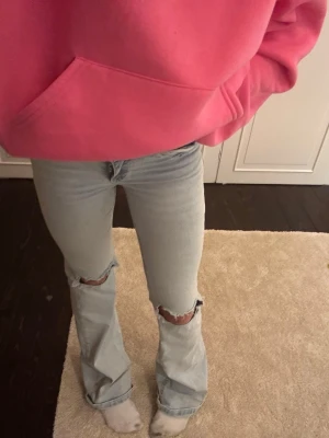 Nelly - Säljer ett par ljusblå bootcut jeans från nelly med slitna hål vid knäna och klassiska röda sömmar på bakfickorna. Jeansen har hög midja och är tillverkade i denim med en skön passform. Perfekta för en avslappnad och trendig look.