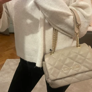 Beige handväska från Valentino - Säljer en snygg beige handväska från Valentino med quiltat mönster och guldfärgad kedjerem. Väskan har en klassisk rektangulär form och logotypen i guld framtill. Perfekt accessoar för att lyfta din outfit.
