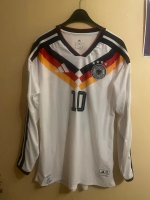 Musiala - Tyskland - VM #10  - Säljer en Tysklands landslagströja, VM-kval, player version, långärmad med Musiala #10. Tröjan är i player issue-utförande med lätt och ventilerande material, figurnära passform och hög kvalitet från adidas. Klassisk vit design med de ikoniska färgdetaljerna över bröstet och DFB-emblemet. Mycket fint skick, sparsamt använd och väl omhändertagen. Perfekt för samlare, matchspel eller som en exklusiv supportertröja. XL. Men sitter som L