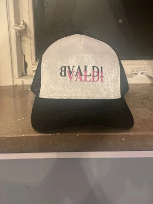 Vit och svart truckerkeps från Valdi - Säljer en stilren truckerkeps från Valdi med vit frontpanel och svart mesh på sidorna och bak. Kepsen har svart böjd skärm och broderad text i svart och rosa på framsidan. Perfekt för en avslappnad streetwear-look.