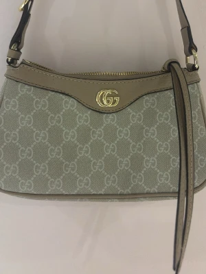 Gucci beige axelremsväska GG canvas - Snygg beige handväska från Gucci med läderdetaljer och guldfärgad logga framtill. Väskan har en axelrem i läder och dragkedja upptill. Säljer pågrund av för stor kollektion , helt ny skick 9/10