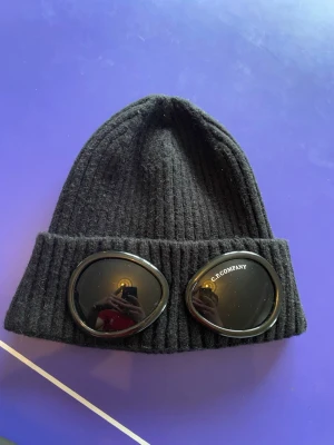 Svart mössa med goggles från C.P. Company - Säljer en svart ribbstickad mössa från C.P. Company med ikoniska inbyggda goggles framtill. Mössan är tillverkad i mjukt material och har en uppvikt kant. Perfekt för dig som vill sticka ut med en streetwear-inspirerad look.