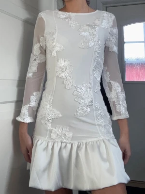 Vit klänning  - Snygg vit kortklänning med transparenta långärmar och vackra applikationer i blommigt mönster. Klänningen har volang nedtill och en feminin, figurnära passform. Perfekt för student. Aldrig använd men har tyvärr inte prislappen kvar