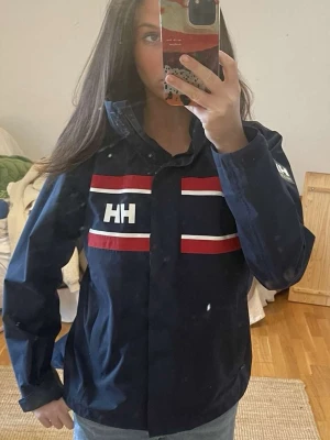 Helly Hansen jacka  - Snygg marinblå vindjacka från Helly Hansen med röda och vita ränder över bröstet och HH-logga på bröstet och ärmen. Jackan har hög krage och dragkedja framtill, perfekt för blåsiga dagar. Klassisk sportig look som funkar året runt.