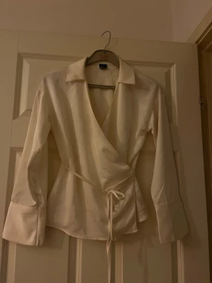 Krämvit omlottblus med krage - Snygg krämvit omlottblus med knytband i midjan och klassisk krage. Blusen har långa ärmar och en figurnära passform som ger en stilren look. Perfekt att styla med jeans eller kjol för en trendig outfit.