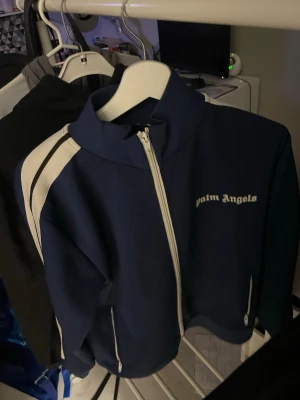 Blå zip hoodie från Palm Angels - Snygg blå zip hoodie från Palm Angels med vita detaljer längs ärmarna och vit dragkedja. Märkets logga syns på bröstet. Tillverkad i Italien och har en sportig look med hög krage och fickor med dragkedja. Perfekt för dig som gillar streetwear.