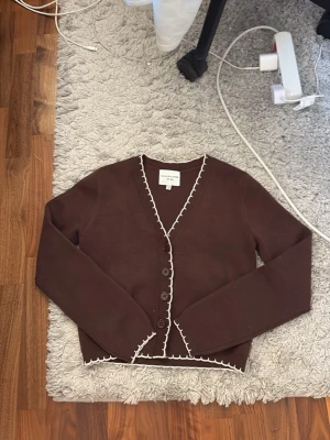 Brun kofta  - Mysig brun kofta med v-ringning och dekorativa vita sömmar längs kanterna. Koftan har långa ärmar, knäppning framtill med fem knappar och är något croppad i modellen. Perfekt att slänga över en topp.