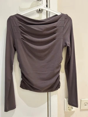  Ny Mörkgrå långärmad topp med drapering - Snygg mörkgrå långärmad topp med draperad effekt över bröstet och axlarna. Tillverkad i mjukt, stretchigt material som sitter skönt på kroppen. Perfekt för dig som gillar stilrena plagg med en twist.