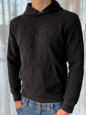 Svart Ralph Lauren hoodie med svart logga, storlek S - Svart Ralph Lauren hoodie med svart broderat tryck på magen  58% polyester 42% bomull  Mycket bra skick, inga snören annars jättefräsch   Storlek S                                                                   Axelbredd: 46 cm Längd: 64 cm