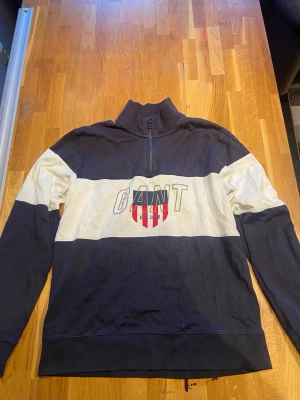 Blå och vit halfzip hoodie från GANT - Snygg blå och vit hoodie från GANT med halfzip och hög krage. Stor GANT-logga och röd sköld på bröstet. Tillverkad i mjukt bomullsmaterial och har breda färgblock. Perfekt för en avslappnad och sportig stil.
