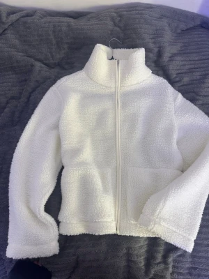 Vit teddyjacka med dragkedja - Mysig vit teddyjacka i fluffigt material med hög krage och hel dragkedja framtill. Jackan har två stora fickor på framsidan och en avslappnad passform. Perfekt för dig som gillar en enkel men trendig look.