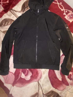 Svart Jack & Jones jacka  - Snygg svart Jack & Jones jacka med dragkedja framtill och två fickor på sidorna. Modellen har huva med dragsko och ribbade muddar vid ärmslut och nederkant. Perfekt för en avslappnad och stilren look.