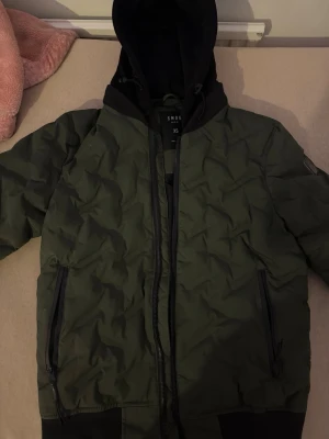 Olivgrön bomberjacka från SMOG - Snygg olivgrön bomberjacka från SMOG med svart huva och dragkedja framtill. Jackan har två sidofickor med dragkedja och ribbade muddar vid ärmslut och nederkant. Materialet är troligtvis polyester och jackan har en modern, avslappnad passform. Den är väldigt fin men lite stickat där vid luvan. Pris kan diskuteras