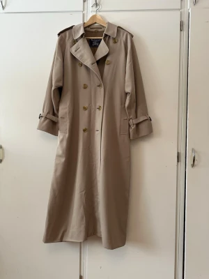 Burberry Trenchcoat. 100% Original  - Lång beige trenchcoat med klassisk dubbelknäppning, axelklaffar och bälte i midjan. Jackan har bred krage, spännen vid ärmsluten och en stilren siluett. Perfekt för dig som gillar en tidlös och clean look. Materialet ser ut att vara bomull eller bomullsblandning.