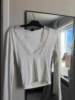 Vit långärmad topp med spets - Snygg vit långärmad topp med djup v-ringning och spetsdetaljer längs urringningen. Modellen är ” figurnära ”! toppen passar bra till vardags om man vill styla upp outfiten lite eller om man ska till nåt finare tillfälle passar den med! man kan även vända toppen bak och fram så spetsen kommer ner i ryggen. Det är jättefint! 