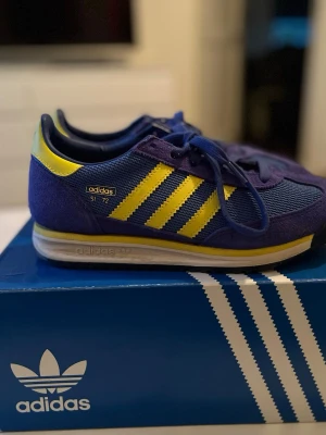 Adidas SL 72 blå/gula sneakers - Snygga Adidas SL 72 sneakers i blått med gula detaljer och klassiska tre ränder på sidan. Ovandelen är i mesh och mocka, med svart yttersula och gula inslag på hälen och sulan. Retroinspirerad design med låg profil och snörning.