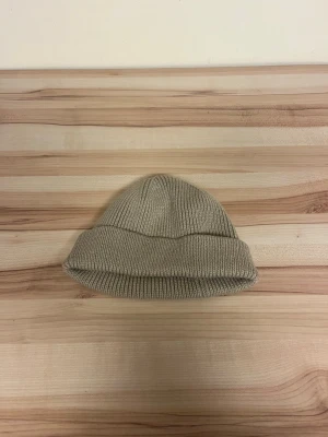 Beige stickad mössa  - Snygg beige stickad mössa från i modellen Short Fisher Beanie. Mössan är ribbstickad och har uppvikt kant. Tillverkad i 94% akryl och 6% elastan för en mjuk och stretchig känsla. Perfekt för dig som gillar enkel och stilren design. Aldrig använd!
