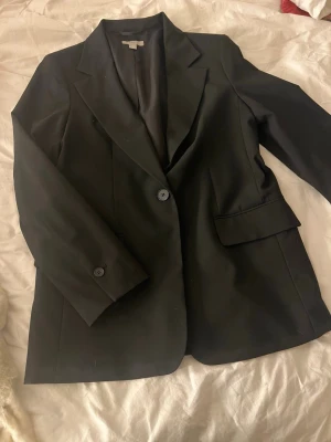 Svart kavaj från H&M, storlek S - Stilren svart kavaj från H&M med klassisk passform och två knappar framtill. Kavajen har två fickor med lock och smala slag. Perfekt för en clean och snygg look. Materialet känns mjukt och följsamt.
