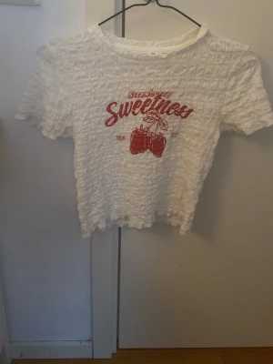 Vit topp med röd text och jordgubbar - Söt vit topp med struktur och korta ärmar. Framsidan har röd text 'Strawberry Sweetness' och en jordgubbsillustration. Toppen är croppad och har en ribbad känsla i materialet. Perfekt för dig som gillar gulliga detaljer och somrig stil.