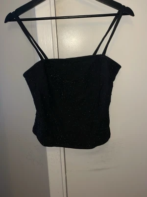 Svart glitter linne med smala band - Snygg svart linnetopp med tunna axelband och diskret glittrigt tyg. Toppen har en croppad passform och raka linjer, perfekt för att styla med höga jeans eller kjol. Inga synliga loggor eller märken.