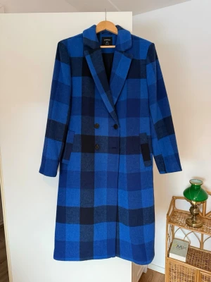Blårutig ullkappa från Lindex - Snygg dubbelknäppt kappa från Lindex i blå och svart rutig design. Kappan har klassisk krage, raka ärmar och två fickor framtill. Tillverkad i ullblandning för extra värme och stil. Perfekt för dig som vill sticka ut med färg i höst och vinter.