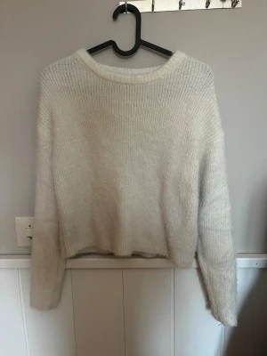 Vit fluffig stickad tröja - Mysig vit stickad tröja med fluffig känsla och rund halsringning. Tröjan har en rak passform och långa ärmar, perfekt för lager-på-lager. Materialet ser ut att vara en mjuk blandning, troligen med inslag av mohair eller liknande.