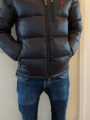 Svart dunjacka från Polo Ralph Lauren - Mörkblå dunjacka från Polo Ralph Lauren med röd logga på bröstet. Jackan har dragkedja framtill, tre fickor med dragkedja och en avtagbar huva. Puffig modell som håller värmen under kalla dagar. Kan säljas billigare vid snabb affär.