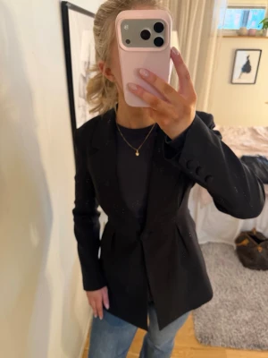 Svart figursydd kavaj - Stilren svart kavaj med figurnära passform och klassiska slag. Kavajen har en enkelknäppning och dekorativa knappar vid ärmsluten. Perfekt att matcha med jeans eller dressade byxor för en snygg look.