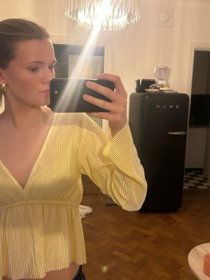 Ljusgul v-ringad blus med volang - Superfin ljusgul blus med v-ringning och volang nertill. Långa ärmar och lätt plisserat tyg ger en luftig känsla. Perfekt för dig som vill ha en söt och trendig topp till våren eller sommaren.