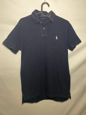 Ralph Lauren Piké  - Marinblå pikétröja från Polo Ralph Lauren i custom slim fit. Klassisk krage med knappar och broderad logga på bröstet. Tillverkad i mjuk bomull och har korta ärmar. Perfekt för en stilren och avslappnad look.