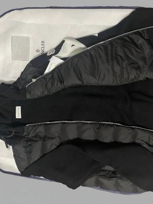 Svart dunjacka från Moncler - Snygg svart dunjacka från Moncler med dragkedja framtill och hög krage. Jackan har Moncler-logga på ärmen och är tillverkad i en kombination av dunfyllda paneler och mjukt tyg.  Pris kan diskuteras vid snabb affär.
